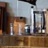 A french press beaker sits on a wooden block in a kitchen as a man pours coffee into it as one of the coffee brew methods for gourmet coffee