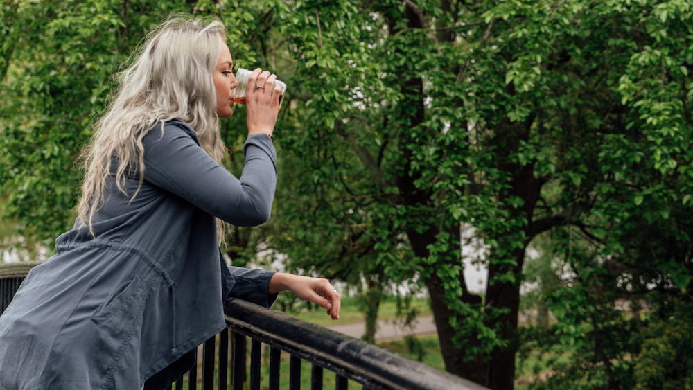 blond woman standing on a bridge with trees in background, drinking one of cooper's cask coffee wine barrel aged coffee choices